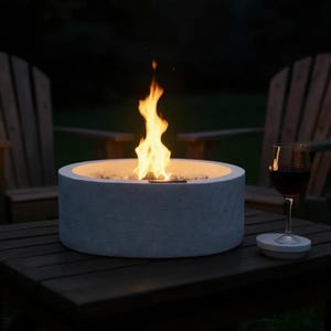 May include: A lit, gray concrete fire pit with small rocks inside, sits on a dark wooden table. A glass of red wine rests on a matching coaster. Two wooden chairs are in the background, with string lights overhead.