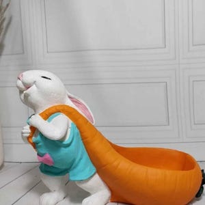 Könnte beinhalten: Eine weiße Hasenfigur, die ein blaues Hemd trägt und einen großen, orangefarbenen, karottenförmigen Pflanzkübel trägt. Der Hase hat rosa Innenohren und lächelt. Der Pflanzkübel ist für kleine Pflanzen oder Dekorationsartikel gedacht.