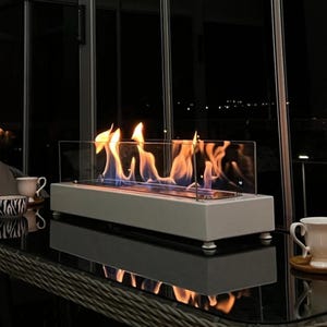 Op de afbeelding: Een moderne, witte tafelhaard met een glazen behuizing, met heldere oranje en blauwe vlammen. De haard is rechthoekig en staat op kleine, afgeronde voetjes. De scène speelt zich binnenshuis af met een donkere achtergrond en een reflecterend oppervlak.