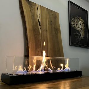 以下が含まれることがあります： 鮮やかな炎を映し出す、ガラスケース付きのモダンな長方形の黒い暖炉。炎は黄色と青の色合いを放っています。暖炉の後ろには、大きな天然木の板と、ヒョウの額縁写真があります。