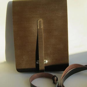 Op de afbeelding: Een rechthoekige bruine leren tas met een verticale riemsluiting en een zilverkleurige metalen knop. De tas is voorzien van een lange, verstelbare schouderriem van bijpassend bruin leer met een metalen gesp en bijbehorende hardware.