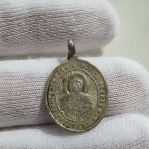 May include: An oval silver-toned metal pendant featuring a relief image of a haloed figure holding a book. The border contains Cyrillic text and a small star symbol. The pendant has a small loop at the top for attachment to a chain or cord.