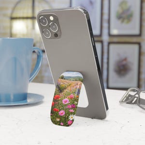 May include: A gray smartphone with a floral pop-up phone grip. The grip features a vibrant field of colorful wildflowers, including red poppies, pink cosmos, and white daisies. The phone is propped up on a white surface.
