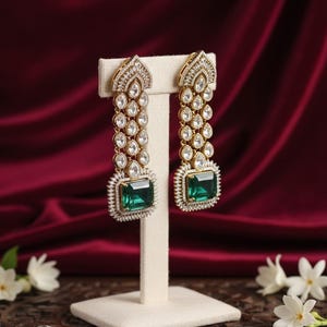 May include: Gold-coloured drop earrings with clear and emerald-green gemstones. The earrings feature a rectangular emerald-green stone at the base, surrounded by smaller clear stones. The design includes a series of clear stones leading to a decorative top.