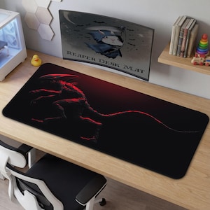 Puede incluir: Alfombrilla de escritorio negra con una ilustración roja y negra de una criatura. La pantalla del monitor en el fondo muestra las palabras "REAPER DESK MAT". Hay un patito de goma amarillo en el escritorio.
