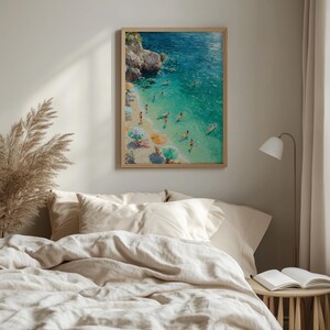 May include: A framed wall art print depicting an aerial view of a coastal beach scene with turquoise water, rocky cliffs, and people swimming or relaxing under colourful umbrellas on the sandy shore, rendered in an impressionistic painting style.
