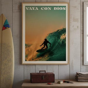 Può includere: Poster incorniciato con la scritta "VAYA CON DIOS" sopra un surfista che cavalca un'onda. Il poster ha una cornice beige ed è appeso a una parete di legno. Una tavola da surf è appoggiata alla parete a sinistra.