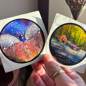 May include: Two round stickers featuring watercolor art. One depicts a figure with wings against a starry sky and fiery landscape. The other shows a person in water with a forest background. The artist's signature is visible.