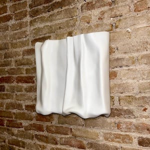 Op de afbeelding: Een witte, abstracte muursculptuur met een gedrapeerd stoffen uiterlijk. De sculptuur is bevestigd aan een bakstenen muur, wat een contrast cre&euml;ert tussen het gladde, witte oppervlak en de ruwe, getextureerde baksteen.