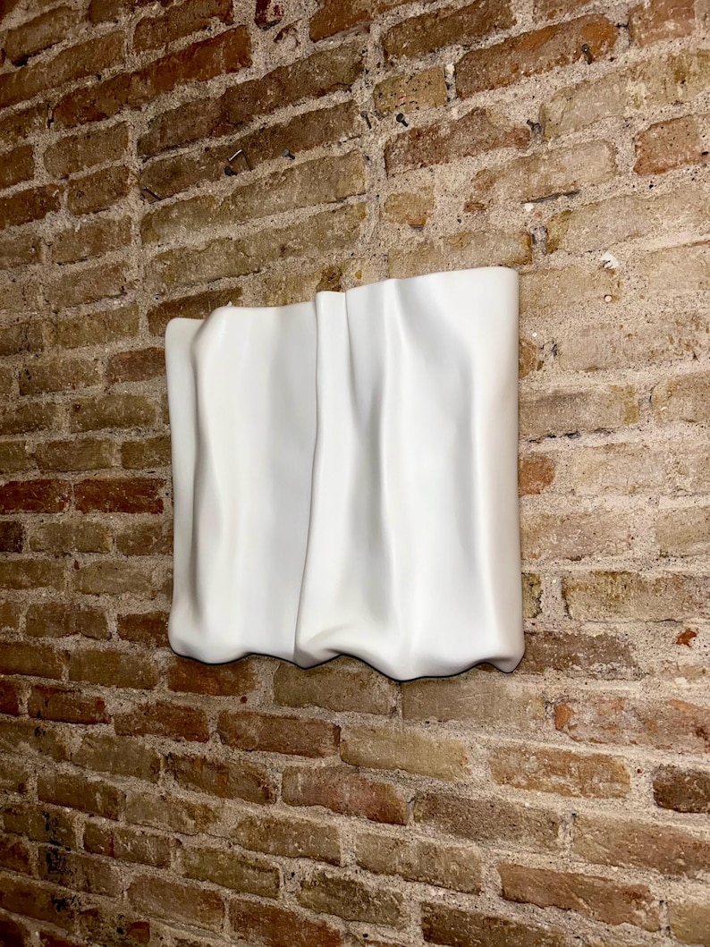 Op de afbeelding: Een witte, abstracte muursculptuur met een gedrapeerd stoffen uiterlijk. De sculptuur is bevestigd aan een bakstenen muur, wat een contrast cre&euml;ert tussen het gladde, witte oppervlak en de ruwe, getextureerde baksteen.