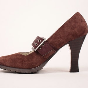 May include: A brown suede Mary Jane style high heel shoe with a buckle closure. The shoe has a lug sole and a dark brown heel. The strap features a silver buckle and a brown elastic band.