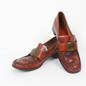 May include: A pair of brown leather loafers with a gold buckle accent. The loafers have a pointed toe and a low heel.
