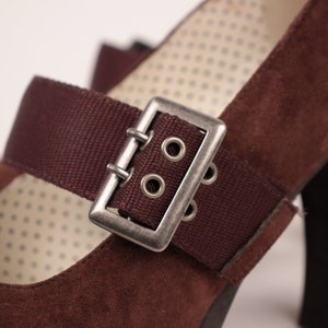 May include: Close-up of a brown suede Mary Jane heel shoe. The shoe features a dark brown strap with a silver buckle. The interior lining has a dotted pattern. The heel is a dark brown color.