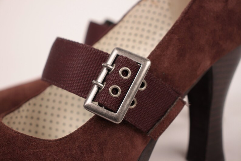 May include: Close-up of a brown suede Mary Jane heel shoe. The shoe features a dark brown strap with a silver buckle. The interior lining has a dotted pattern. The heel is a dark brown color.