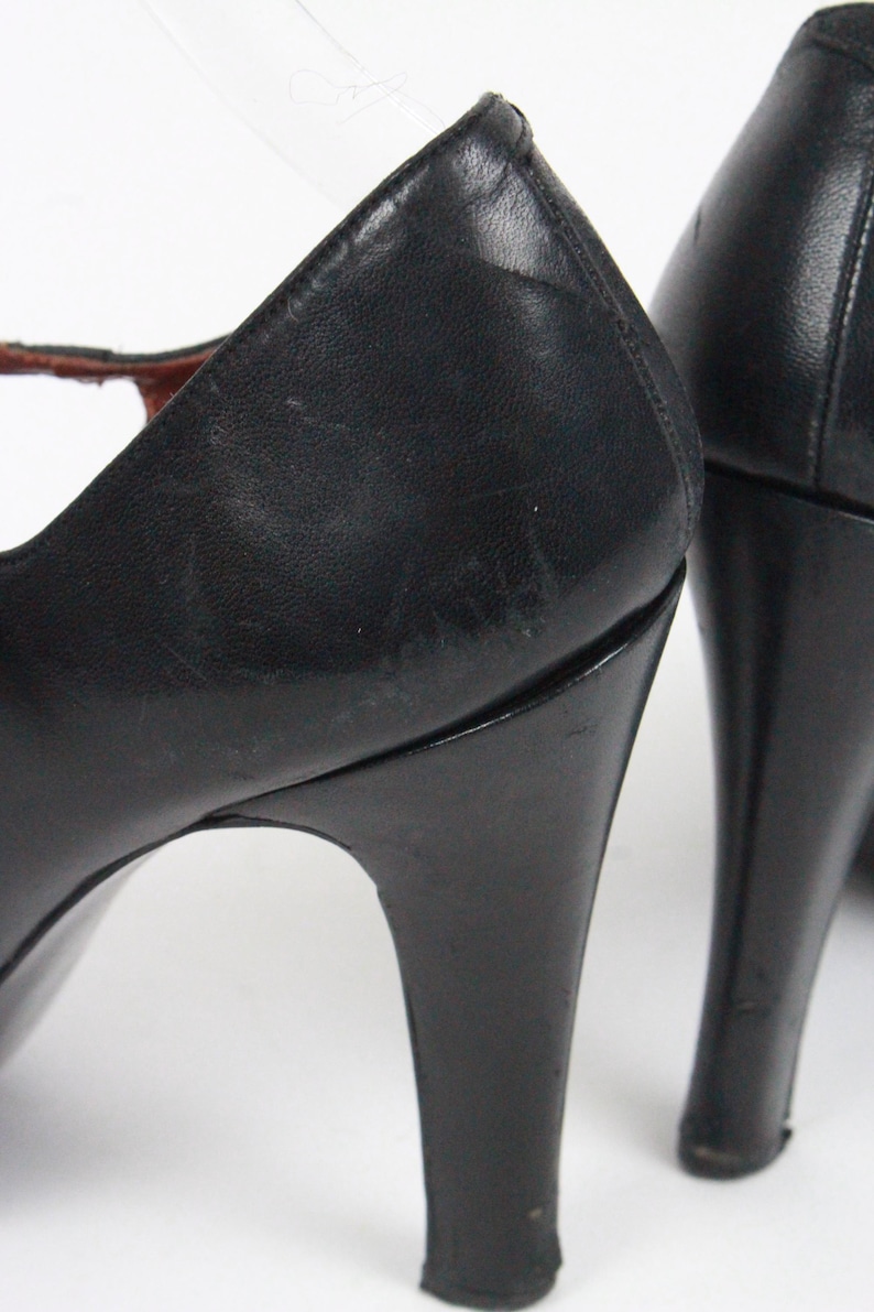 May include: Black leather high-heeled shoes. The shoes have a classic design with a rounded toe and a slender heel. The heels are approximately 3 inches tall. The shoes appear to be vintage.