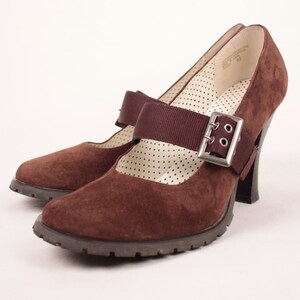 May include: Pair of brown suede Mary Jane heels with a strap and silver buckle. The shoes have a chunky heel and a textured sole. The interior features a white polka dot pattern.