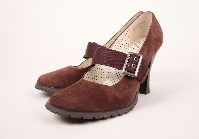 May include: Pair of brown suede Mary Jane heels with a strap and silver buckle. The shoes have a chunky heel and a textured sole. The interior features a white polka dot pattern.