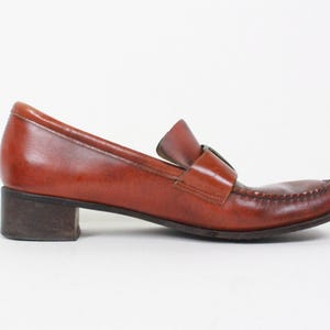 May include: Brown leather heeled loafers with a buckle detail. The loafers have a low block heel and a pointed toe.