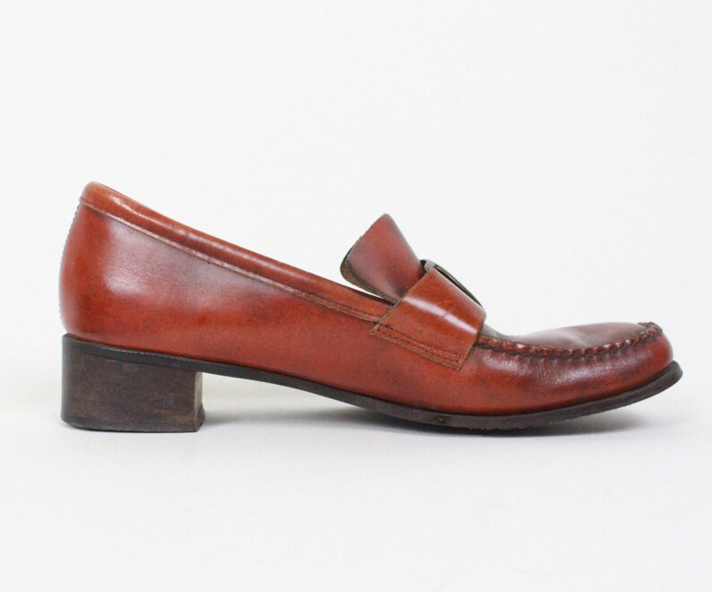 May include: Brown leather heeled loafers with a buckle detail. The loafers have a low block heel and a pointed toe.