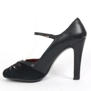 May include: Black high-heeled shoe with a Mary Jane strap. The shoe features a rounded toe with cut-out details and a high heel. The upper part of the shoe is made of black leather and suede.