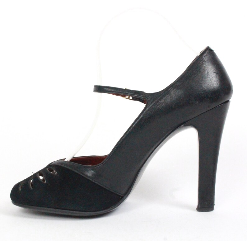 May include: Black high-heeled shoe with a Mary Jane strap. The shoe features a rounded toe with cut-out details and a high heel. The upper part of the shoe is made of black leather and suede.