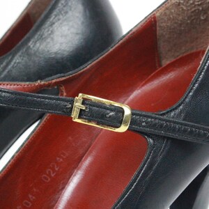 May include: Close-up of a pair of black leather Mary Jane heels. The shoes feature a strap with a gold-colored buckle and a red insole. The heels are high and the shoes appear to be vintage.