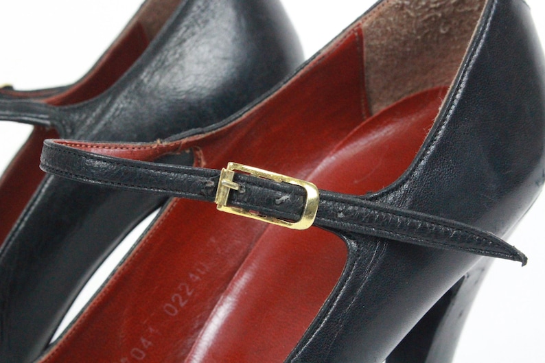 May include: Close-up of a pair of black leather Mary Jane heels. The shoes feature a strap with a gold-colored buckle and a red insole. The heels are high and the shoes appear to be vintage.