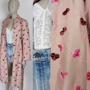 May include: A light pink long coat with a mandarin collar, adorned with pink and burgundy floral embellishments. The coat is open, revealing a white lace top and denim shorts. The overall style is vintage and feminine.