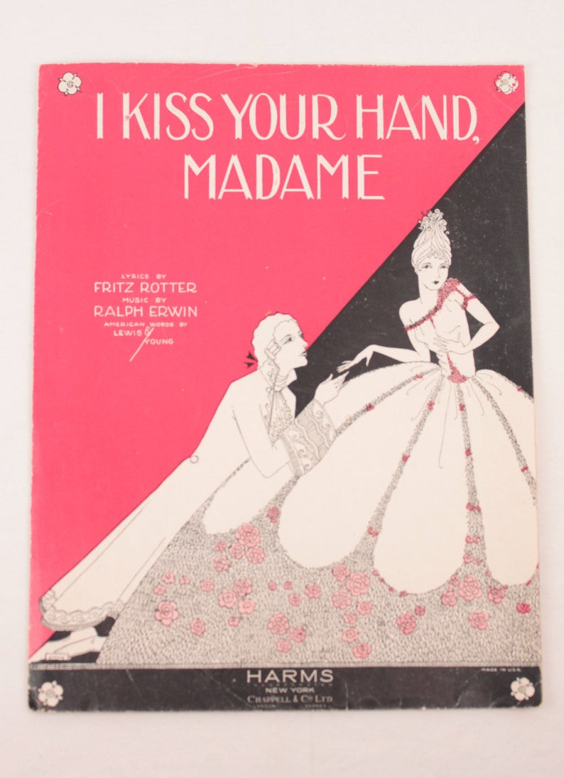 May include: A vintage sheet music cover with a pink and black background. The cover features a black and white illustration of a man and woman in a romantic setting. The title of the song is "I Kiss Your Hand, Madame".