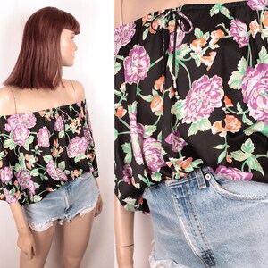 May include: A black off-the-shoulder top with a floral print in shades of pink, green, and orange. The top has a gathered neckline and a loose fit.