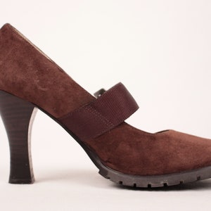 May include: Brown suede Mary Jane heel with a strap across the top of the foot. The heel is dark brown and flared. The sole is a dark brown, lugged style. The shoe is set against a white background.