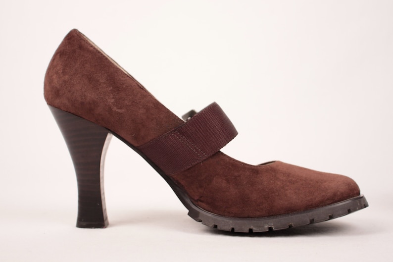 May include: Brown suede Mary Jane heel with a strap across the top of the foot. The heel is dark brown and flared. The sole is a dark brown, lugged style. The shoe is set against a white background.