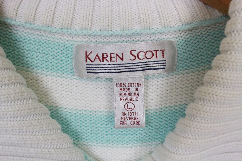 Op de afbeelding: Een wit en lichtblauwe gestreepte trui met een wit label waarop "KAREN SCOTT" en "100% KATOEN GEMAAKT IN DOMINICAANSE REPUBLIEK L RN 13711 ACHTERZIJDE VOOR ZORG" staat.