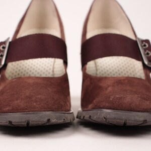 May include: Pair of brown suede Mary Jane style shoes with a strap and buckle closure. The shoes have a dark brown rubber sole and a cushioned insole. The strap is a darker shade of brown. The shoes are displayed on a white background.