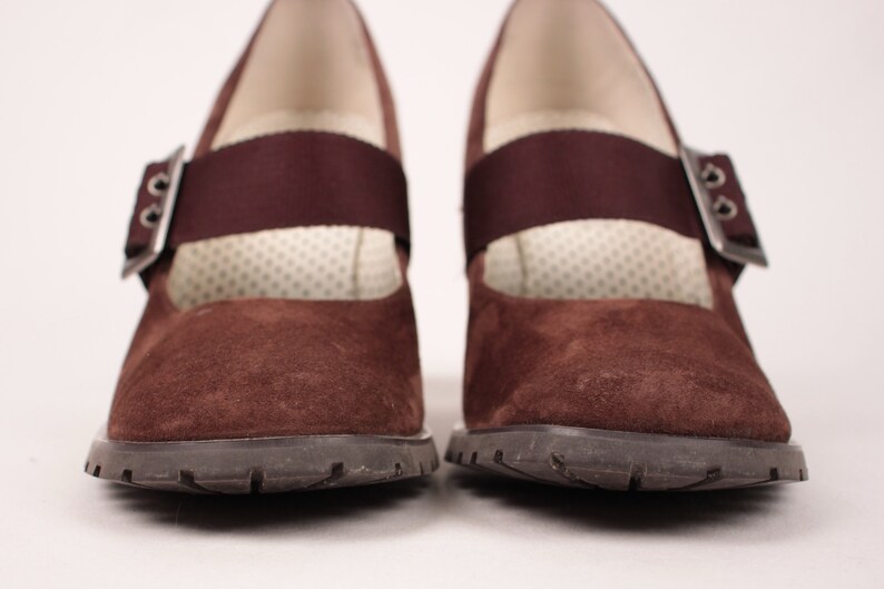 May include: Pair of brown suede Mary Jane style shoes with a strap and buckle closure. The shoes have a dark brown rubber sole and a cushioned insole. The strap is a darker shade of brown. The shoes are displayed on a white background.