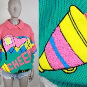 May include: A pink, blue, green, and yellow knit sweater with a cheerleader theme. The sweater features a megaphone, a flag, and the word "CHEER" in large letters.