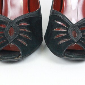 May include: Pair of black suede peep-toe heels. The shoes feature a cut-out design on the vamp and a leather trim. The interior is red. The heels are set against a white background.