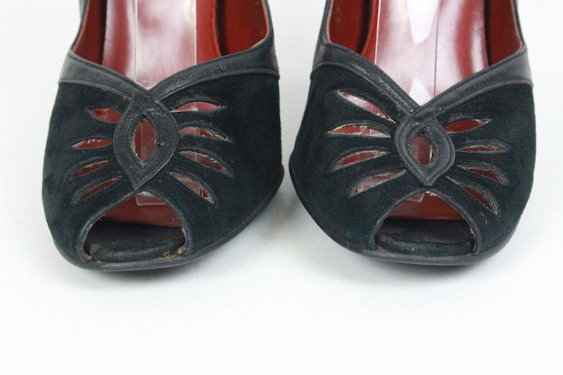 May include: Pair of black suede peep-toe heels. The shoes feature a cut-out design on the vamp and a leather trim. The interior is red. The heels are set against a white background.