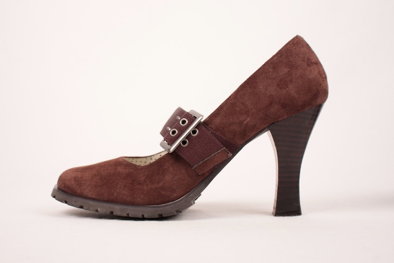 May include: A brown suede Mary Jane style high heel shoe with a buckle closure. The shoe has a lug sole and a dark brown heel. The strap features a silver buckle and a brown elastic band.