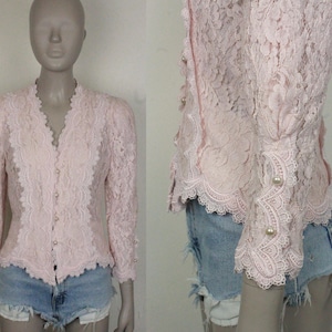 May include: A light pink lace blouse with a scalloped neckline and long sleeves. The blouse features pearl-like buttons and delicate lace detailing. The sleeves are gathered at the shoulders and cuffs. The blouse is paired with denim shorts.