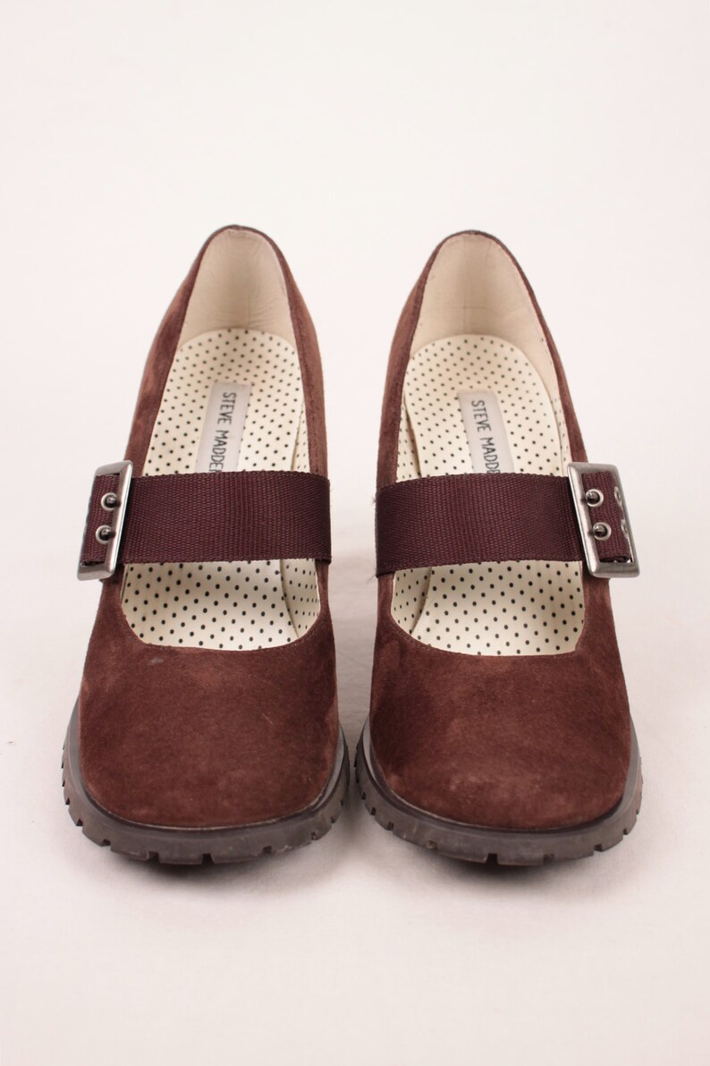 May include: Pair of brown suede Mary Jane style shoes with a strap and buckle closure. The shoes have a chunky sole and a polka dot patterned insole. The brand name "STEVE MADDEN" is printed on the insole.