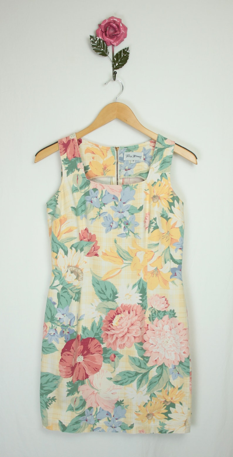 May include: A sleeveless yellow floral dress with a square neckline. The dress has a variety of flowers in shades of pink, red, blue, and yellow. The dress is made by the brand "Paris Blues".