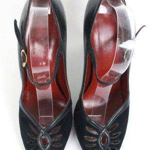 May include: A pair of black vintage shoes with a peep-toe design. The shoes feature a strap with a gold buckle and a cut-out pattern on the vamp. The interior is a deep red color.