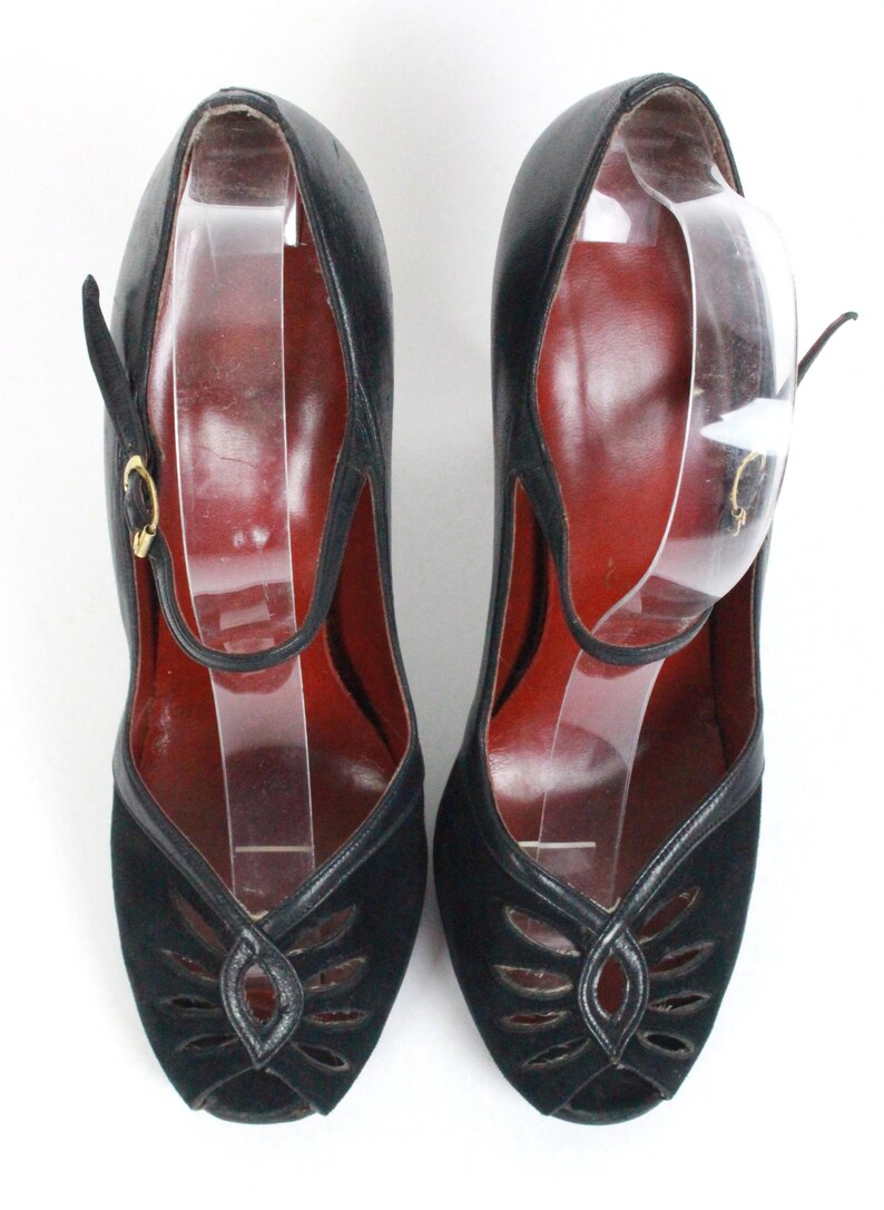May include: A pair of black vintage shoes with a peep-toe design. The shoes feature a strap with a gold buckle and a cut-out pattern on the vamp. The interior is a deep red color.