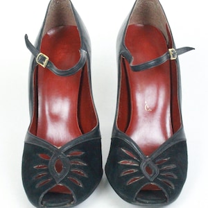 May include: A pair of black peep-toe heels with a strap and buckle closure. The shoes have a black suede design on the front and a red insole. The heels are approximately 3 inches high.