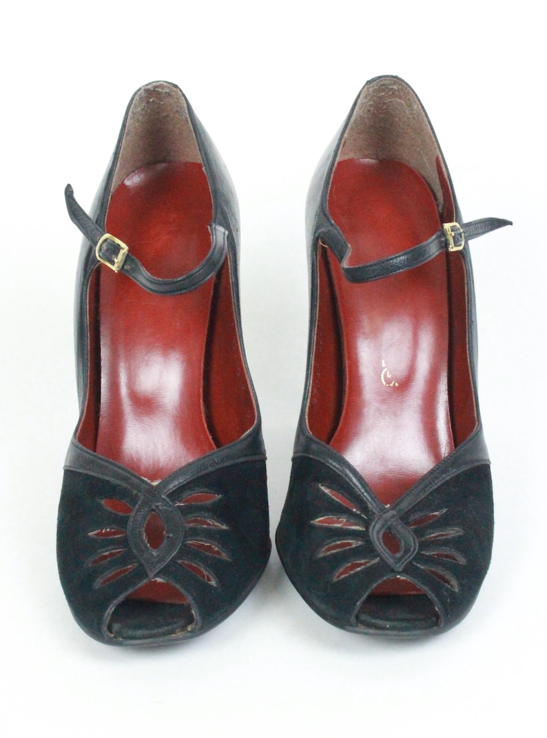 May include: A pair of black peep-toe heels with a strap and buckle closure. The shoes have a black suede design on the front and a red insole. The heels are approximately 3 inches high.