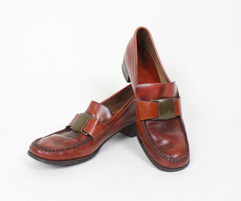 May include: A pair of brown leather loafers with a gold buckle accent. The loafers have a pointed toe and a low heel.