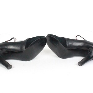 May include: Pair of black leather high-heeled shoes with a strap across the top of the foot. The heels are curved and slender. The shoes are set against a white background.