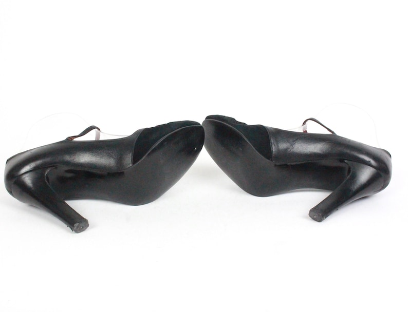 May include: Pair of black leather high-heeled shoes with a strap across the top of the foot. The heels are curved and slender. The shoes are set against a white background.