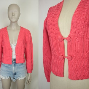 May include: A pink knitted cardigan with a V-neck and button closure. The cardigan has a cable knit pattern and is cropped.